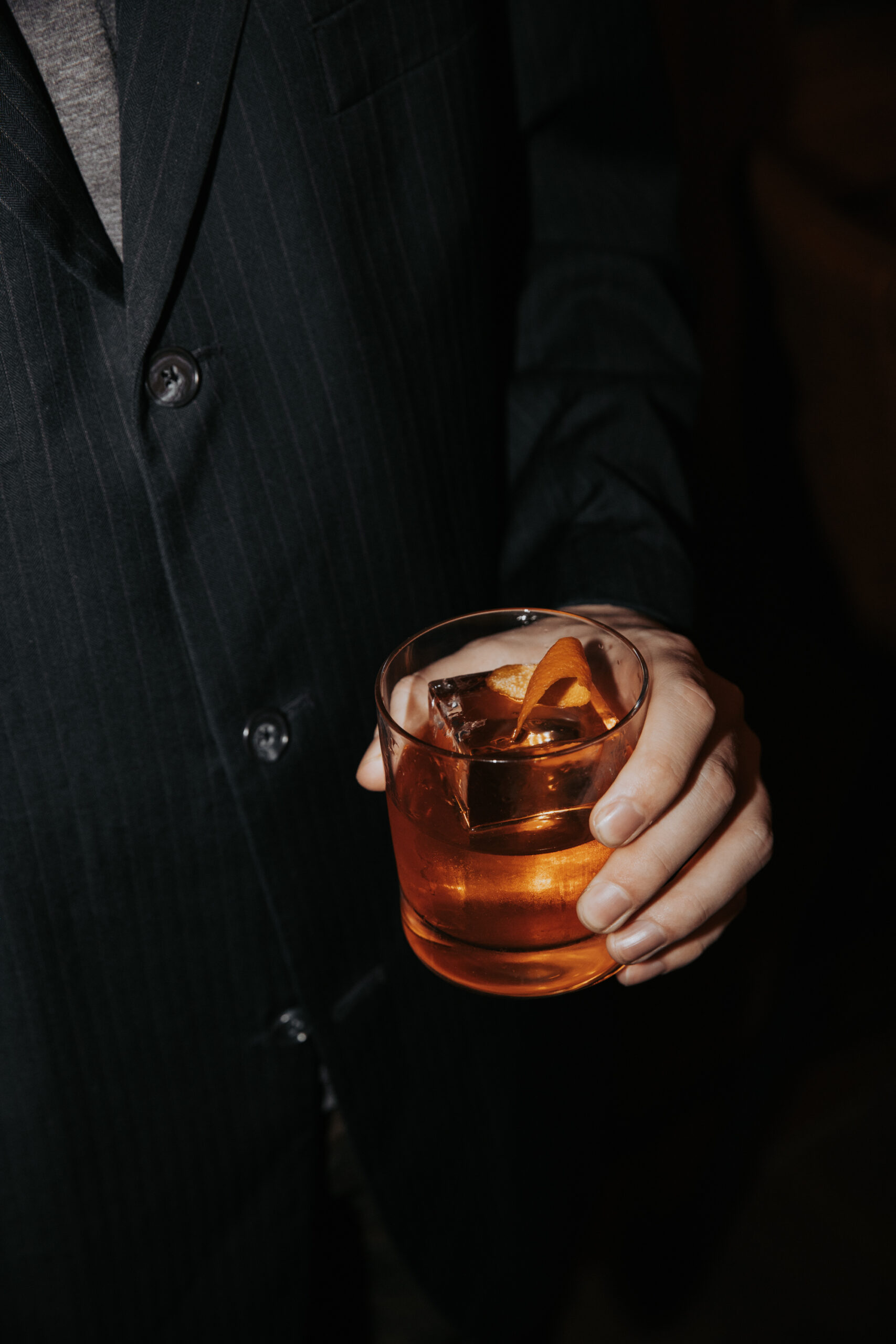 Main d'homme en veston noir tenant un verre de negroni sur fond sombre