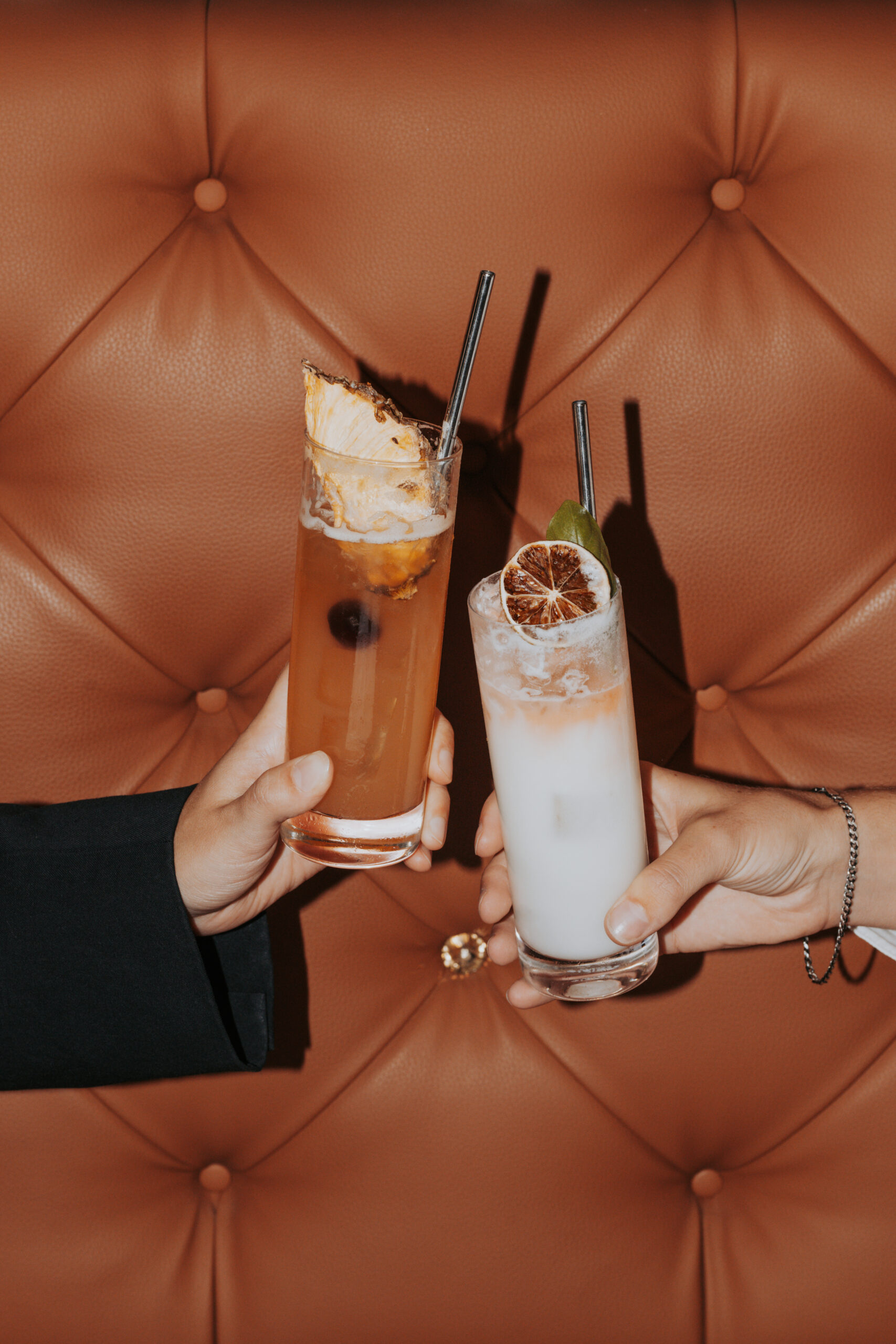 Deux mains trinquant avec des cocktails devant une banquette capitonnée en cuir orange